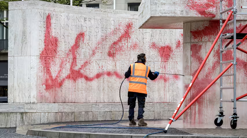 Reiniger in oranje hesje spuit rode verf van het Nationaal Monument op de Dam in Amsterdam nadat het is beklad